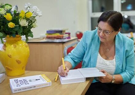 Picture of Carolyn Parry, multi-award-winning career and life coach, author and podcast host of Change Your Story. signing a copy of her book, sitting at a desk with a yellow vast full of spring flowers